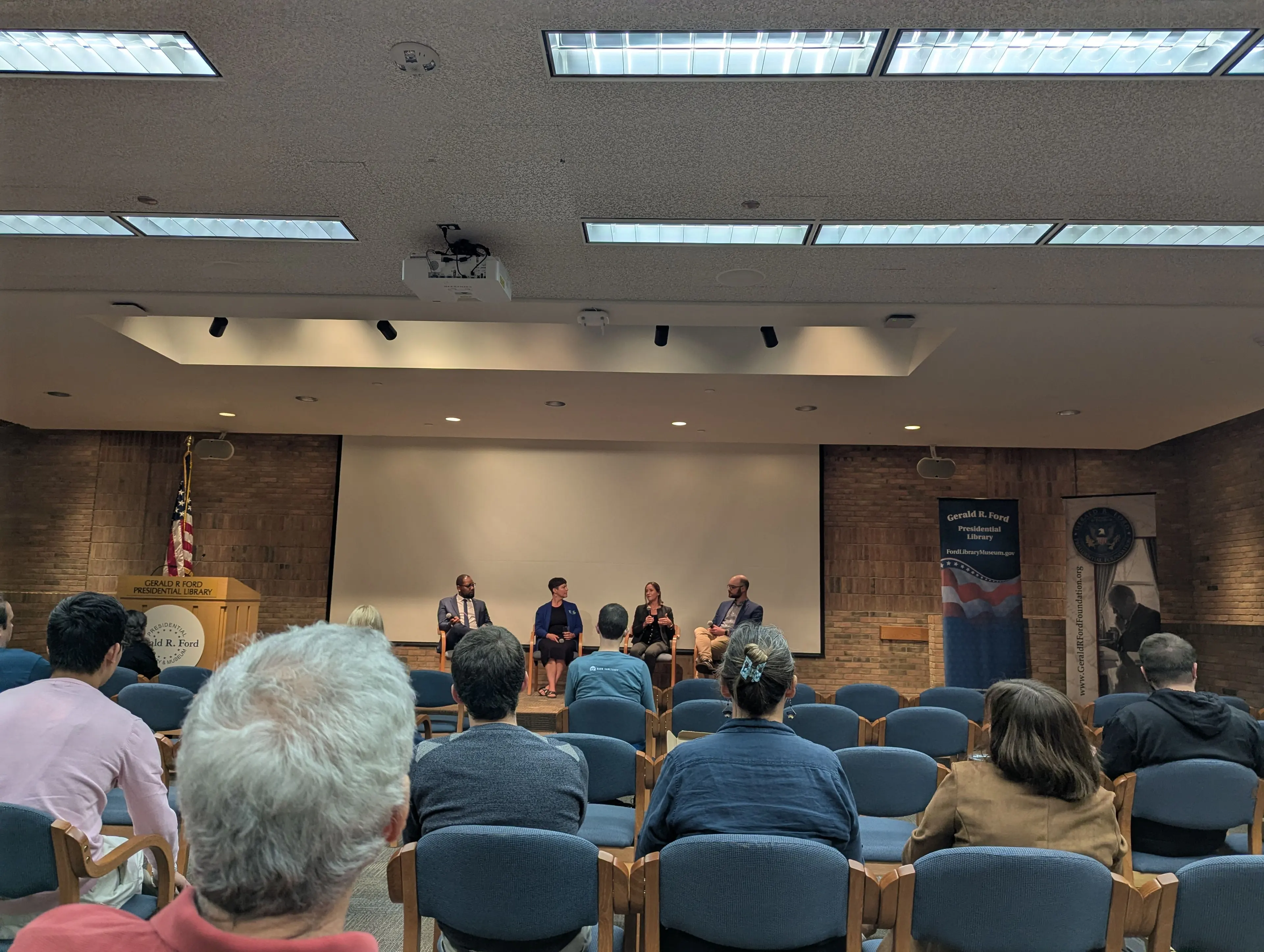 Panelists sit on stage at the Gerald R. Ford Presidential Library during a Climate Week event, speaking to an audience seated in rows of chairs. An American flag and podium are visible to the left, with banners for the library on either side of the stage.
