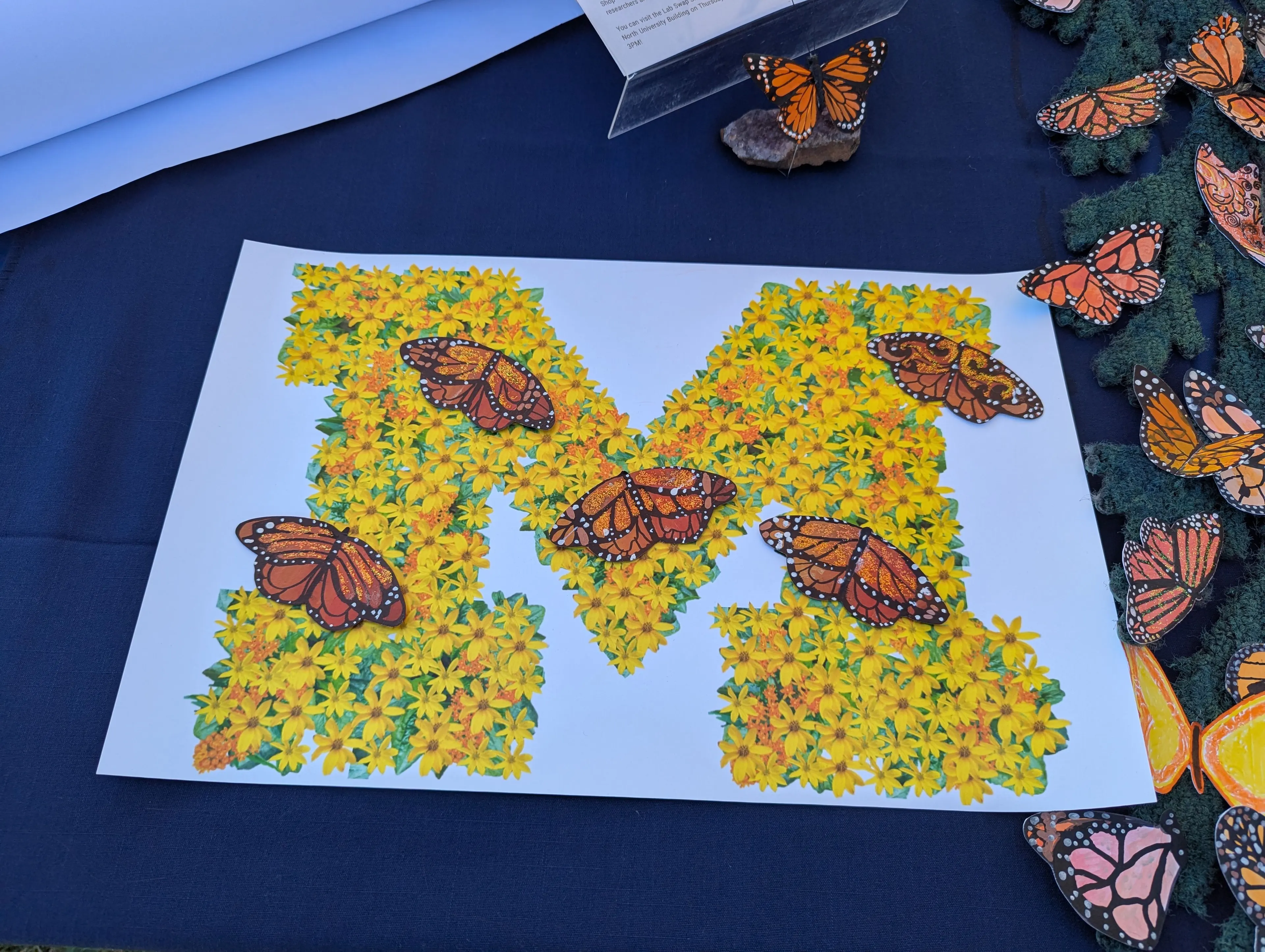 A piece of artwork shaped like the University of Michigan block "M," filled with bright yellow flowers. Several orange-and-black monarch butterflies are placed on top of the flower-filled "M," with additional butterfly cutouts scattered on the surrounding dark blue tablecloth.