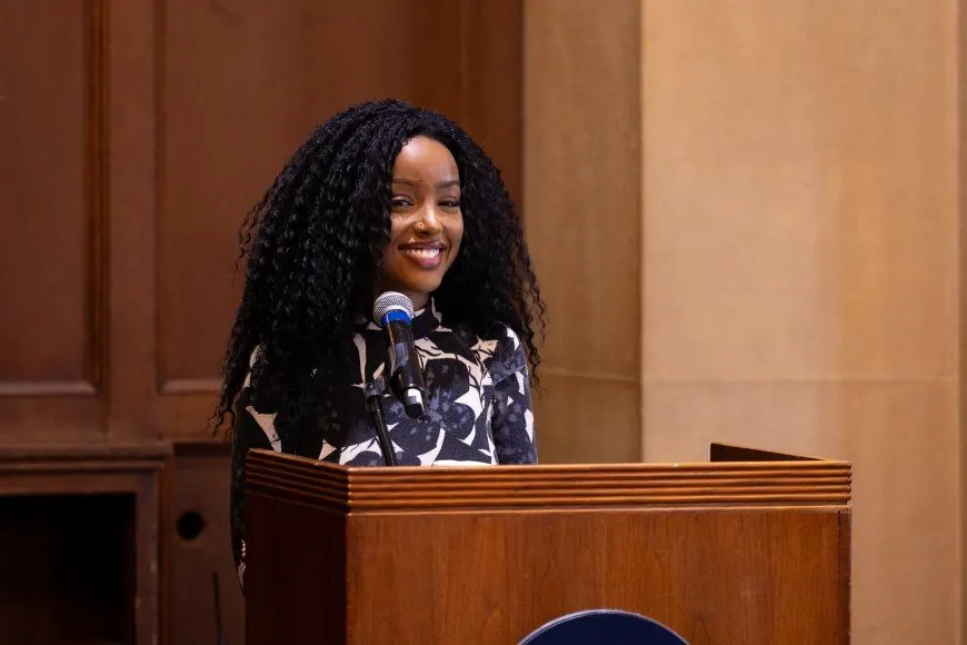 Wawa Gatheru stands at a wooden podium, speaking into a microphone in a formal indoor setting with paneled walls and stone columns.