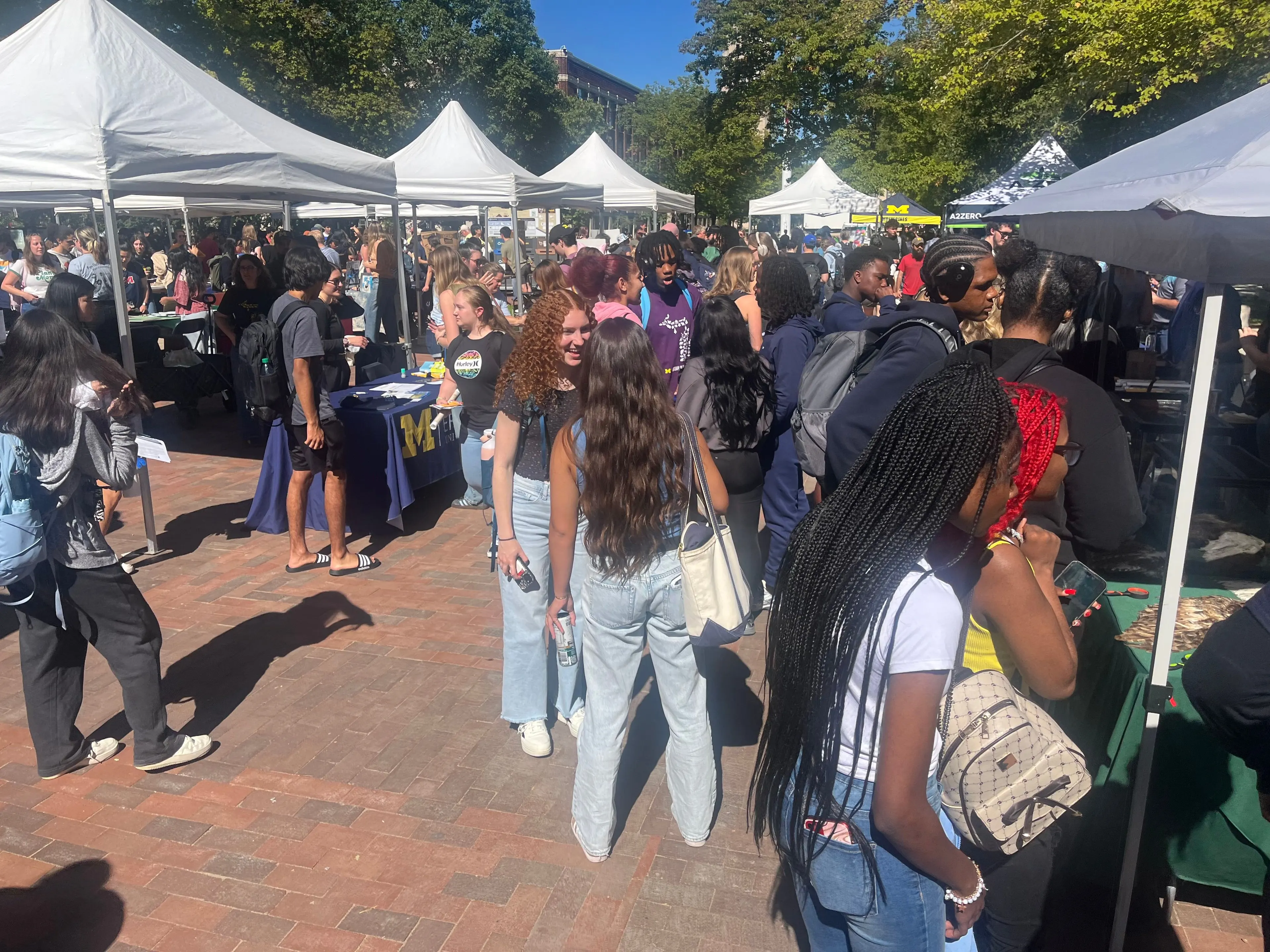 A large crowd of students gathers outdoors under rows of white canopy tents at the University of Michigan’s EarthFest. People are smiling, chatting, and visiting tables with displays about sustainability on a sunny day.