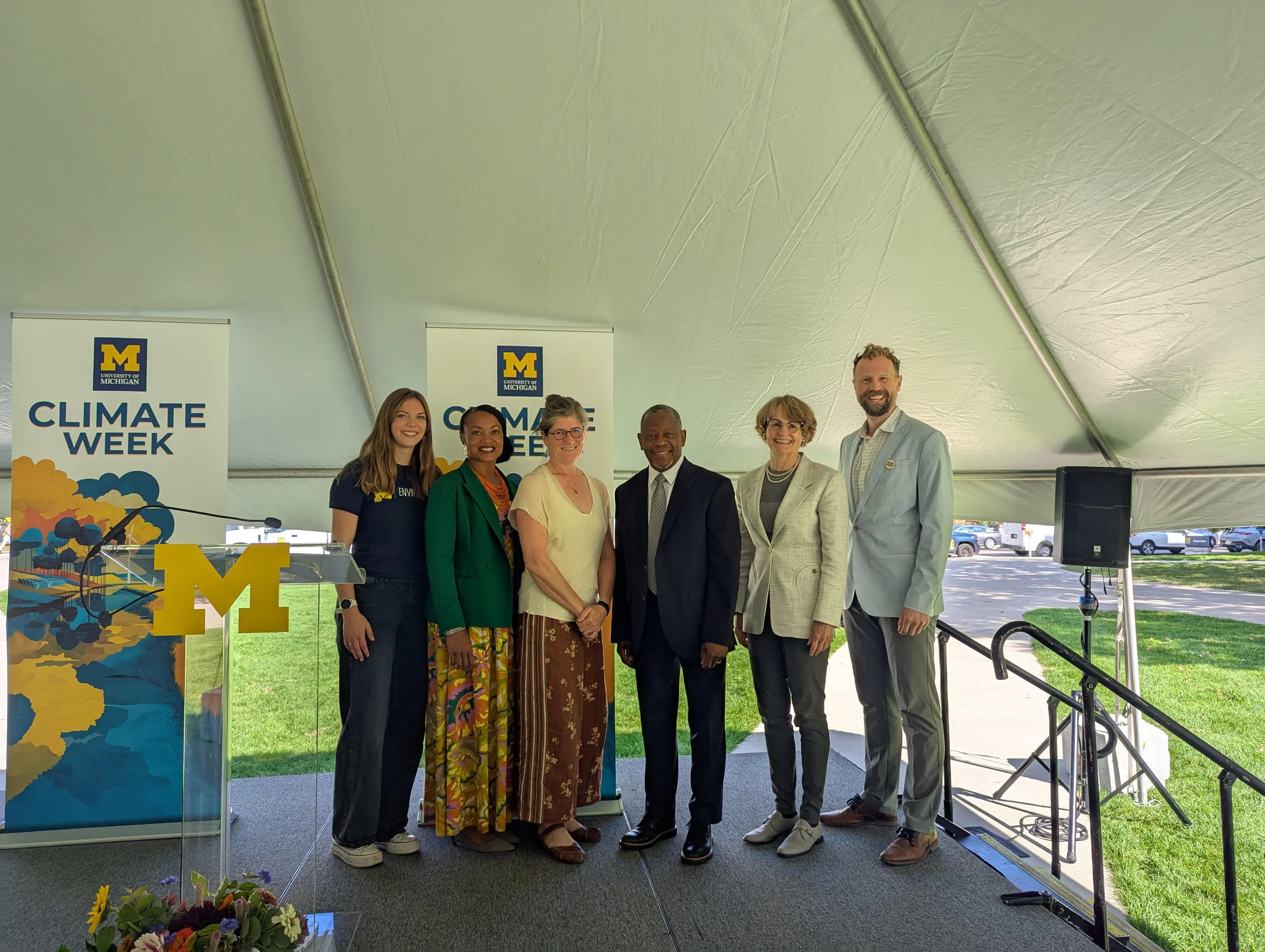 Six people stand together smiling on a stage under a white tent, with University of Michigan “Climate Week” banners and a podium displaying a large yellow block M in the background.
