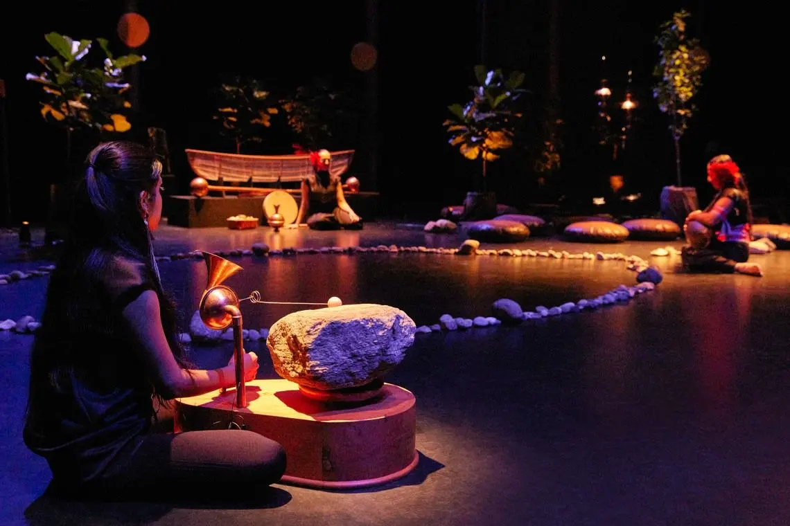 Still from the performance “Nigamon / Tunai” shows three artists seated in a circular arrangement marked by stones, surrounded by plants and warm stage lighting. One performer in the foreground plays an instrument made from a large stone and copper horn, while others sit with drums and ritual objects, creating a contemplative atmosphere that evokes connection between land, sound, and spirit.