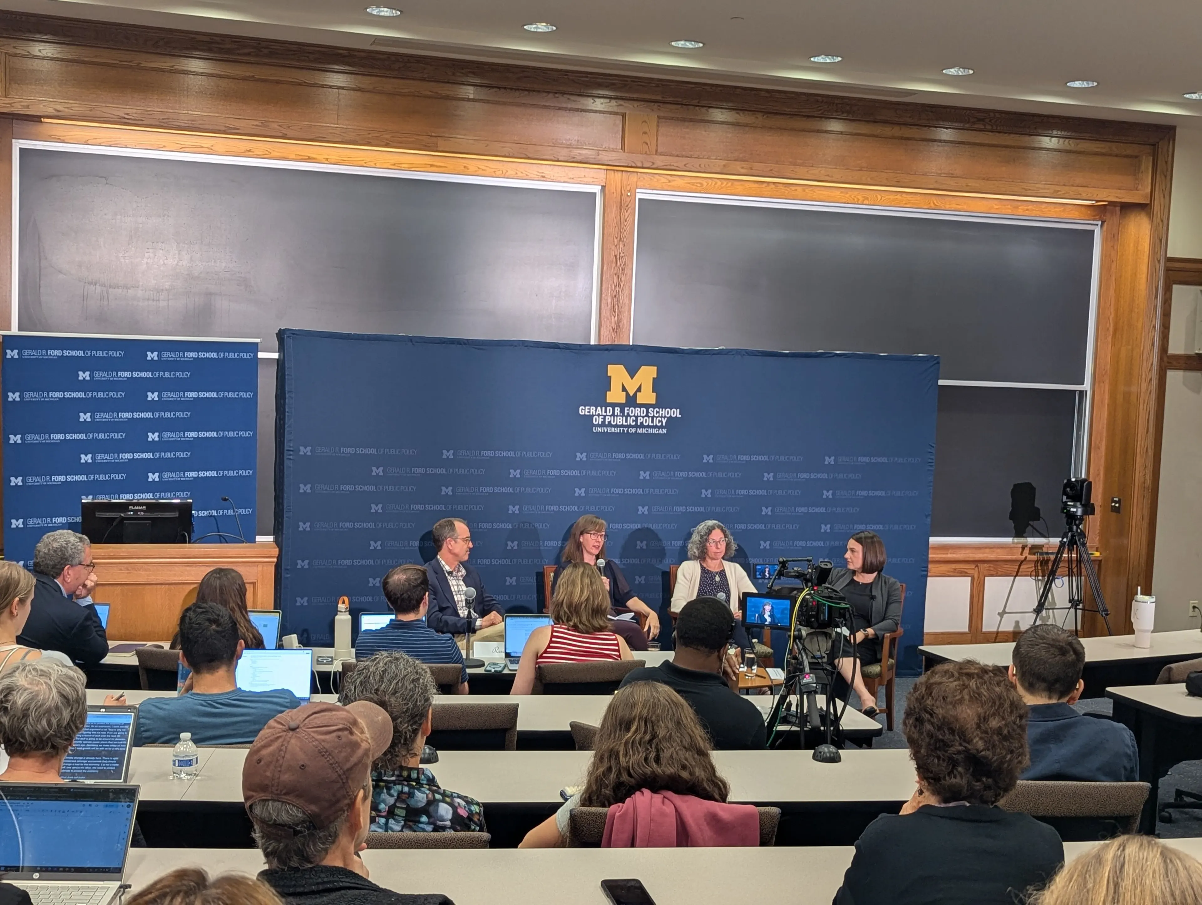 A panel discussion at the University of Michigan’s Gerald R. Ford School of Public Policy features four speakers seated at the front of a lecture hall, with a backdrop displaying the Ford School logo. Audience members fill the room, many taking notes on laptops, while cameras record the event. The discussion appears engaged and interdisciplinary, focusing on energy and climate policy.
